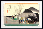 A framed Japanese ukiyo-e woodblock print showing a rural scene with a thatched-roof house, figures near a doorway, bare trees, and a mountain in the background. The print is centered within a white mat and a black frame.
