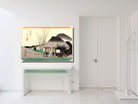 A framed Japanese-style art print depicting a traditional village scene with a thatched-roof house, tree branches, and mountains, displayed on a white wall above a white console table in a bright modern room.