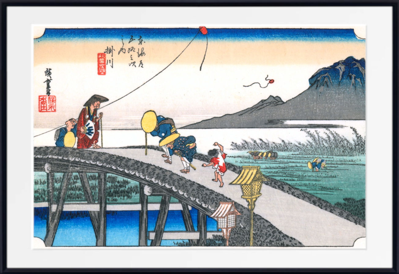 A framed Japanese ukiyo-e woodblock print depicting figures walking across a stone bridge over water, with mountains, reeds, and a pale sky in the background.