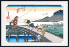 A framed Japanese ukiyo-e woodblock print depicting figures walking across a stone bridge over water, with mountains, reeds, and a pale sky in the background.