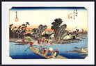 A framed Japanese ukiyo-e woodblock print showing a river scene with a boat, trees, buildings, and a mountain at sunset, presented with a white mat and black frame.