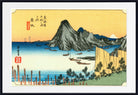 A framed Japanese ukiyo-e woodblock print depicting a coastal mountain scene with boats on blue water, a pale yellow sky, and red seal marks, shown against a white mat and black frame.