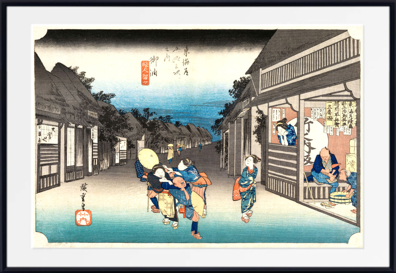 A framed Japanese ukiyo-e woodblock print depicting a street scene with figures walking past traditional buildings under a blue sky, shown with a white mat and black frame.