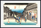 A framed Japanese ukiyo-e woodblock print depicting a street scene with figures walking past traditional buildings under a blue sky, shown with a white mat and black frame.