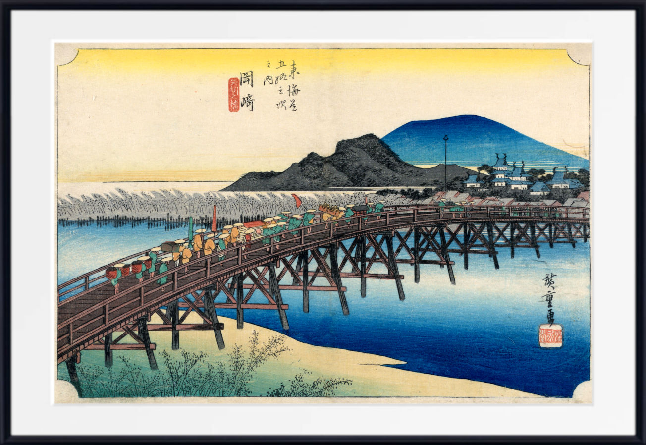 A framed Japanese-style art print depicting a wooden bridge over blue water, with mountains, buildings, and small figures in the scene. The print has a wide white mat and a black frame.