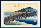 A framed Japanese-style art print depicting a wooden bridge over blue water, with mountains, buildings, and small figures in the scene. The print has a wide white mat and a black frame.