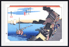 A framed Japanese ukiyo-e woodblock print depicting a coastal harbor scene with boats on blue water, buildings along the shore, and a pale sky with red-orange clouds. The print is shown with a white mat and a black frame.
