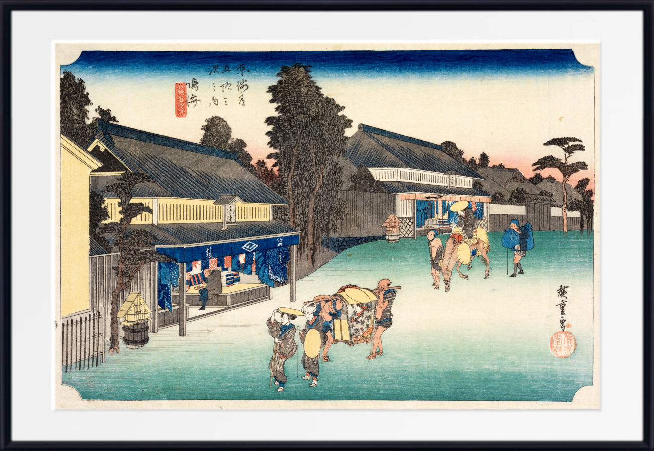 A framed Japanese ukiyo-e woodblock print showing a snowy village street with buildings, trees, and figures, presented with a white mat and black frame.