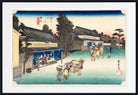 A framed Japanese ukiyo-e woodblock print showing a snowy village street with buildings, trees, and figures, presented with a white mat and black frame.