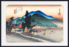 A framed Japanese ukiyo-e woodblock print showing a rural landscape with mountains, trees, houses, and figures in the foreground, presented with a white mat and black frame.