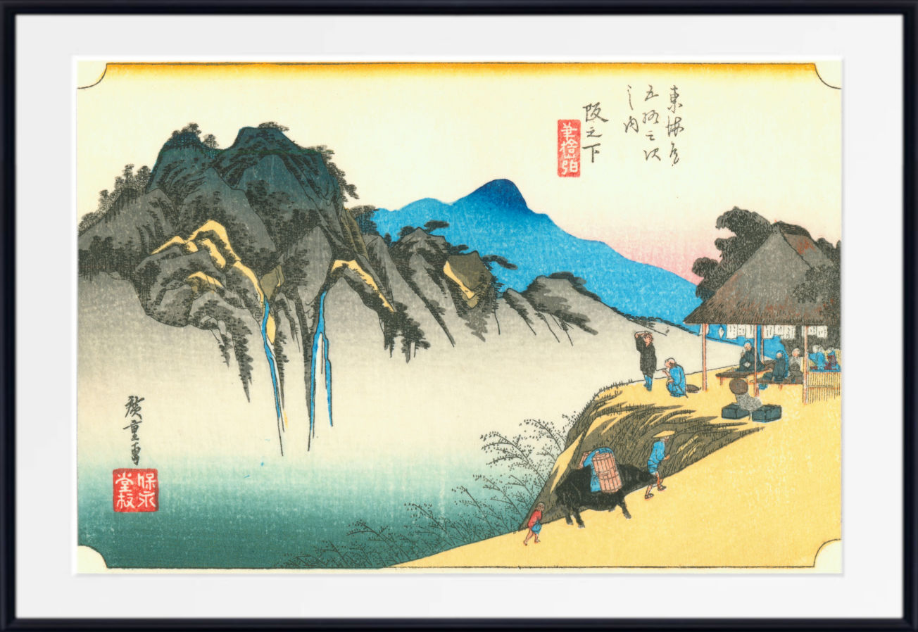 A framed Japanese ukiyo-e woodblock print showing a mountainous landscape with blue mountains, dark rocky cliffs, a pale sky, and figures near a thatched-roof structure on a sandy shore. The print is centered within a white mat and a black frame.