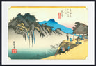 A framed Japanese ukiyo-e woodblock print showing a mountainous landscape with blue mountains, dark rocky cliffs, a pale sky, and figures near a thatched-roof structure on a sandy shore. The print is centered within a white mat and a black frame.