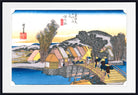 A framed Japanese ukiyo-e woodblock print showing a waterfront village with thatched-roof houses, a wooden bridge, trees, and figures walking under a blue sky. The print is mounted with a white mat and a black frame.