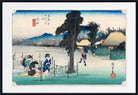 A framed Japanese ukiyo-e woodblock print depicting figures by a body of water with trees, thatched-roof houses, and distant mountains. The artwork is shown with a white mat and a black frame.