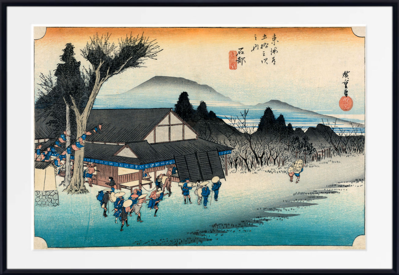 A framed Japanese ukiyo-e woodblock print showing a snowy village scene with figures, a large tree, mountains, and a blue body of water under a pale sky. The print is presented with a white mat and black frame.