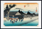 A framed Japanese ukiyo-e woodblock print showing a snowy village scene with figures, a large tree, mountains, and a blue body of water under a pale sky. The print is presented with a white mat and black frame.