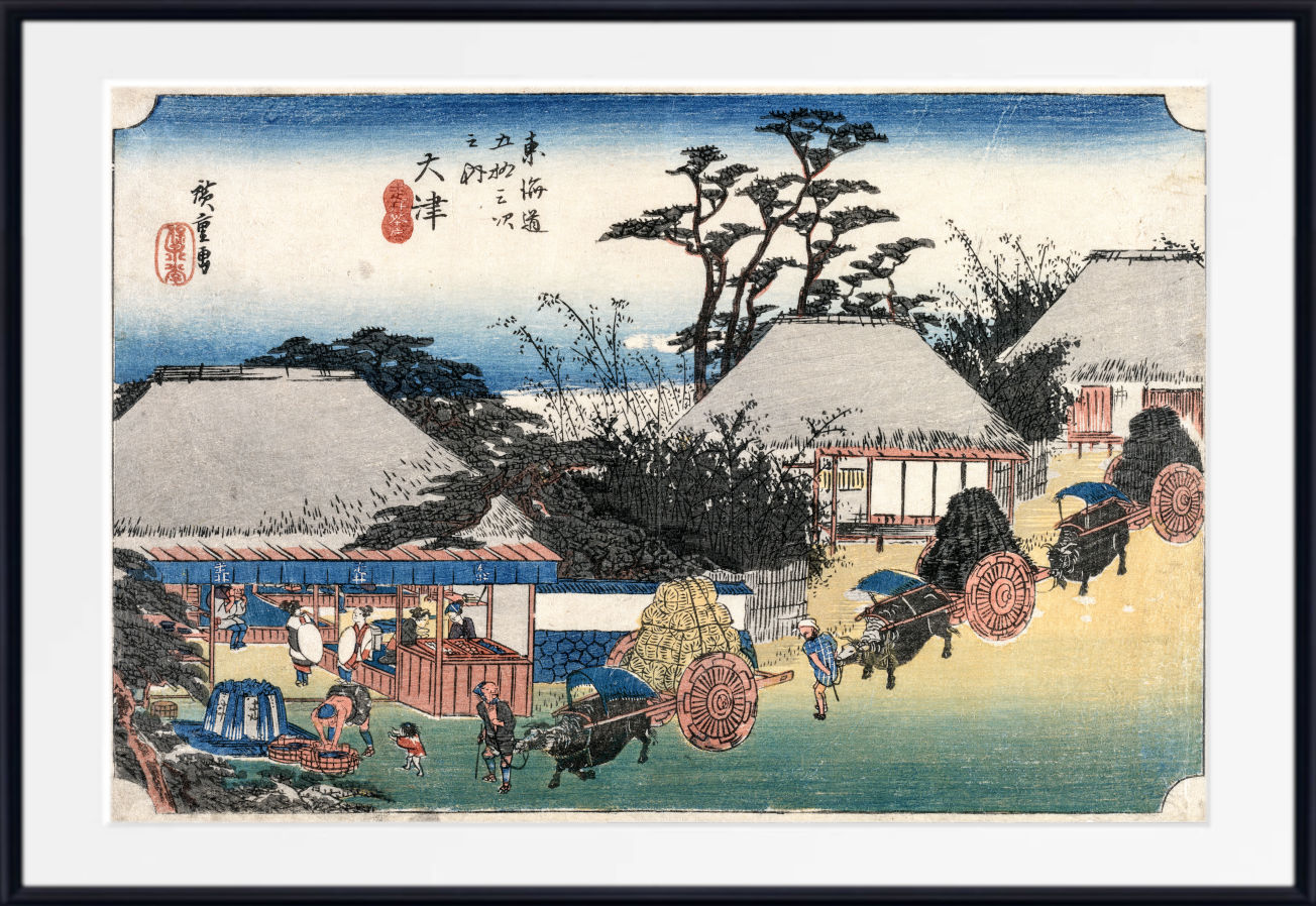 A framed Japanese ukiyo-e woodblock print showing a coastal village scene with thatched-roof houses, figures, boats, and carts, presented with a white mat and black frame.