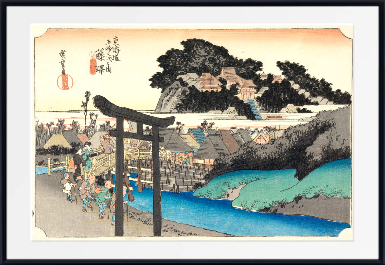 A framed Japanese ukiyo-e woodblock print depicting a coastal village scene with a large dark torii gate in the foreground, blue water, buildings, trees, and a pale sky. The print is shown with a white mat and black frame.