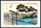 A framed Japanese ukiyo-e woodblock print depicting a coastal village scene with a large dark torii gate in the foreground, blue water, buildings, trees, and a pale sky. The print is shown with a white mat and black frame.