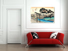 A framed Japanese-style art print depicting a scenic landscape with a torii gate, trees, and blue water, displayed on a white wall above a modern red sofa with black-and-white pillows.