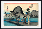 A framed Japanese-style landscape print showing a mountain, water, trees, and two small figures near a post, with red seals and black calligraphy details. The artwork is centered within a white mat and a black frame.