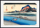 A framed Japanese-style landscape print showing a blue river, sandy shore, reeds, and a mountain with red, orange, and blue tones. The artwork includes black calligraphy and red seals, presented with a white mat and black frame.