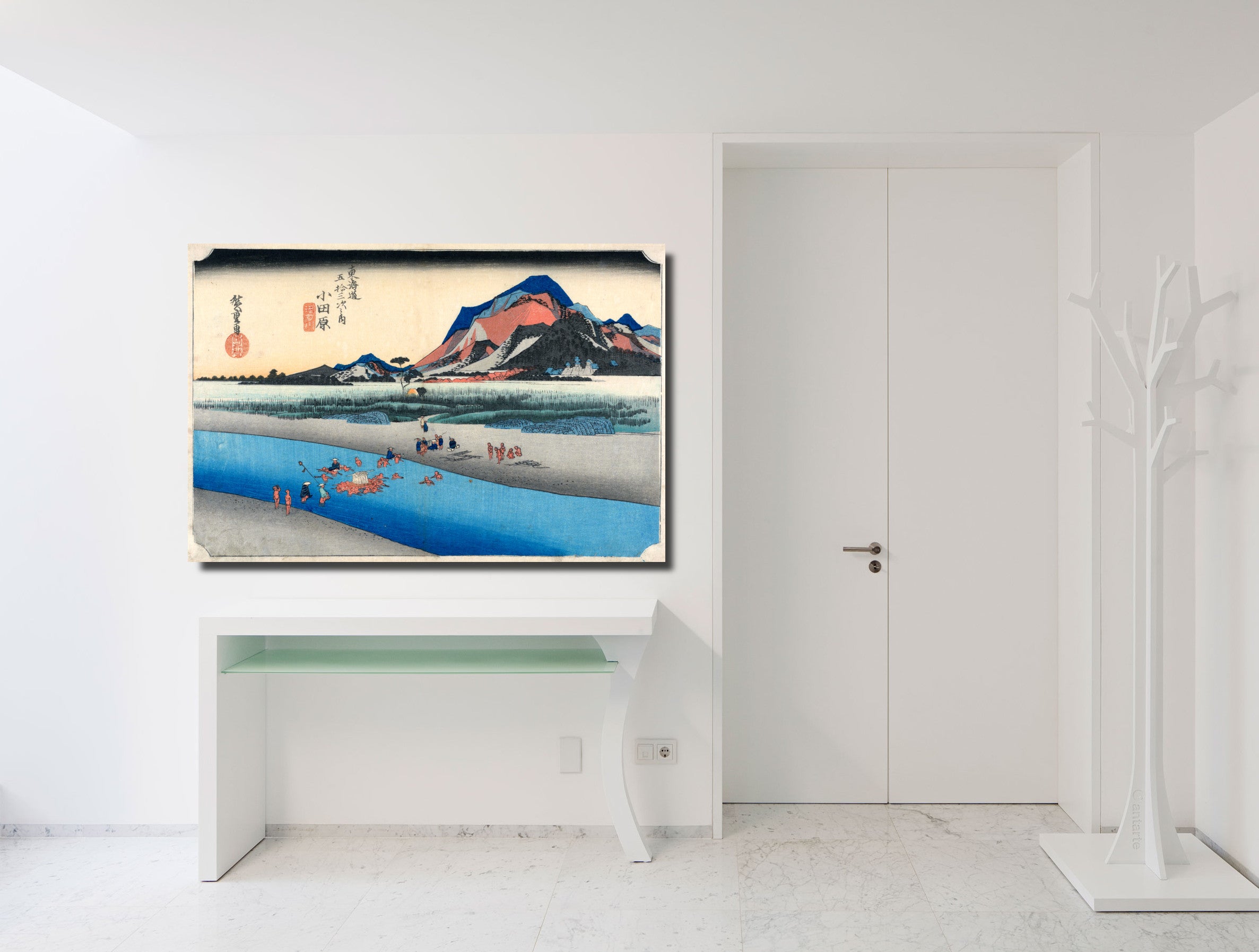 A framed Japanese ukiyo-e style art print depicting a scenic landscape with mountains, water, and figures, displayed on a white wall above a console table in a bright, modern white room.