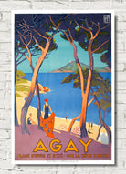A vintage-style travel poster for Agay beach, France, featuring a coastal scene with blue water, sandy shore, trees, and figures near umbrellas, shown hanging against a white brick wall.