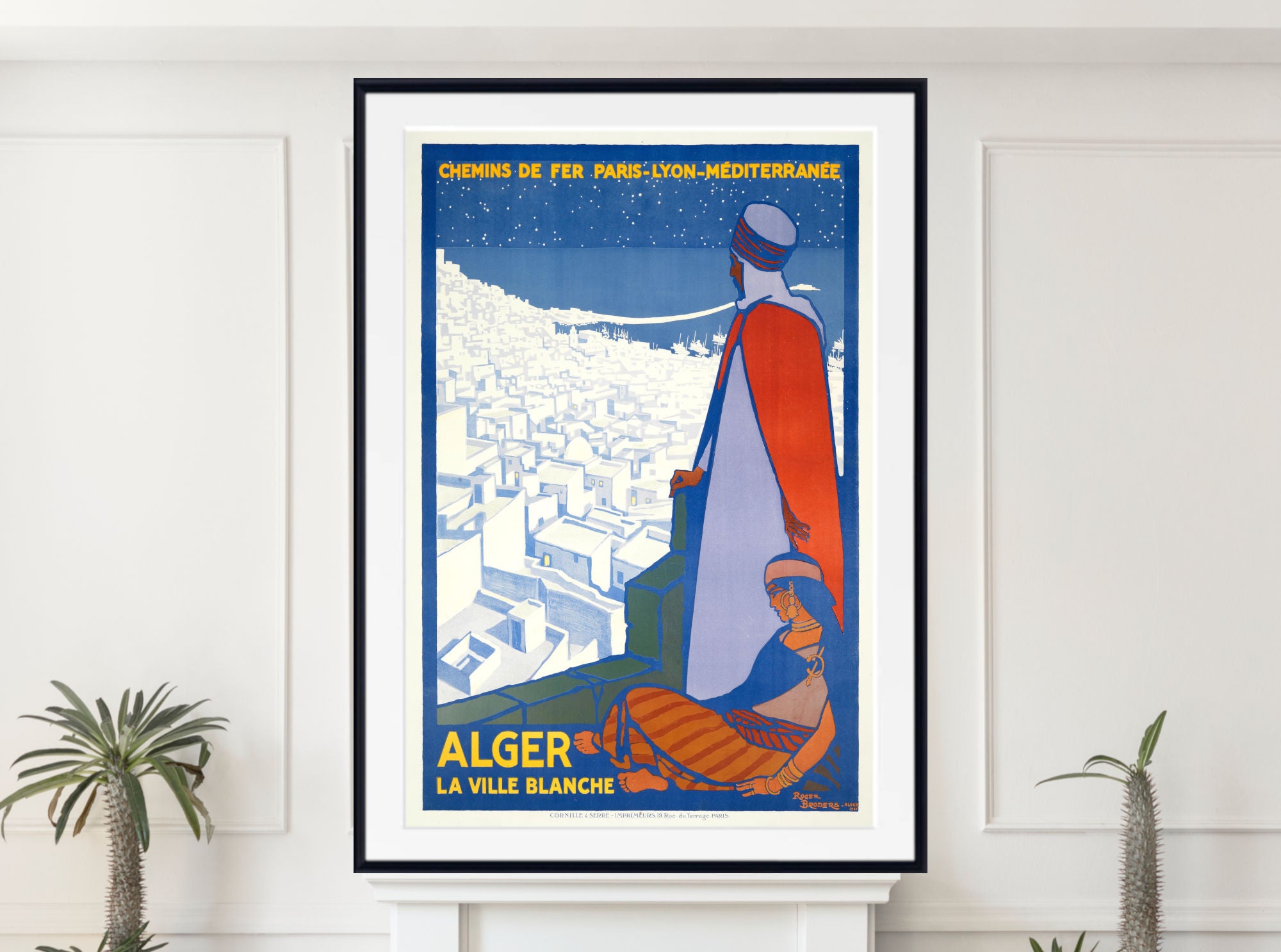A framed vintage-style travel poster of Algér, France, displayed on a white wall above a mantel with small potted plants. The poster features a stylized illustration of a figure in a red and white robe overlooking a white city under a dark blue night sky, with yellow text reading 'CHENIS DE TER PARIS-LYON-MEDITERRANÉE' and 'ALGER LA VILLE BLANCHE'.