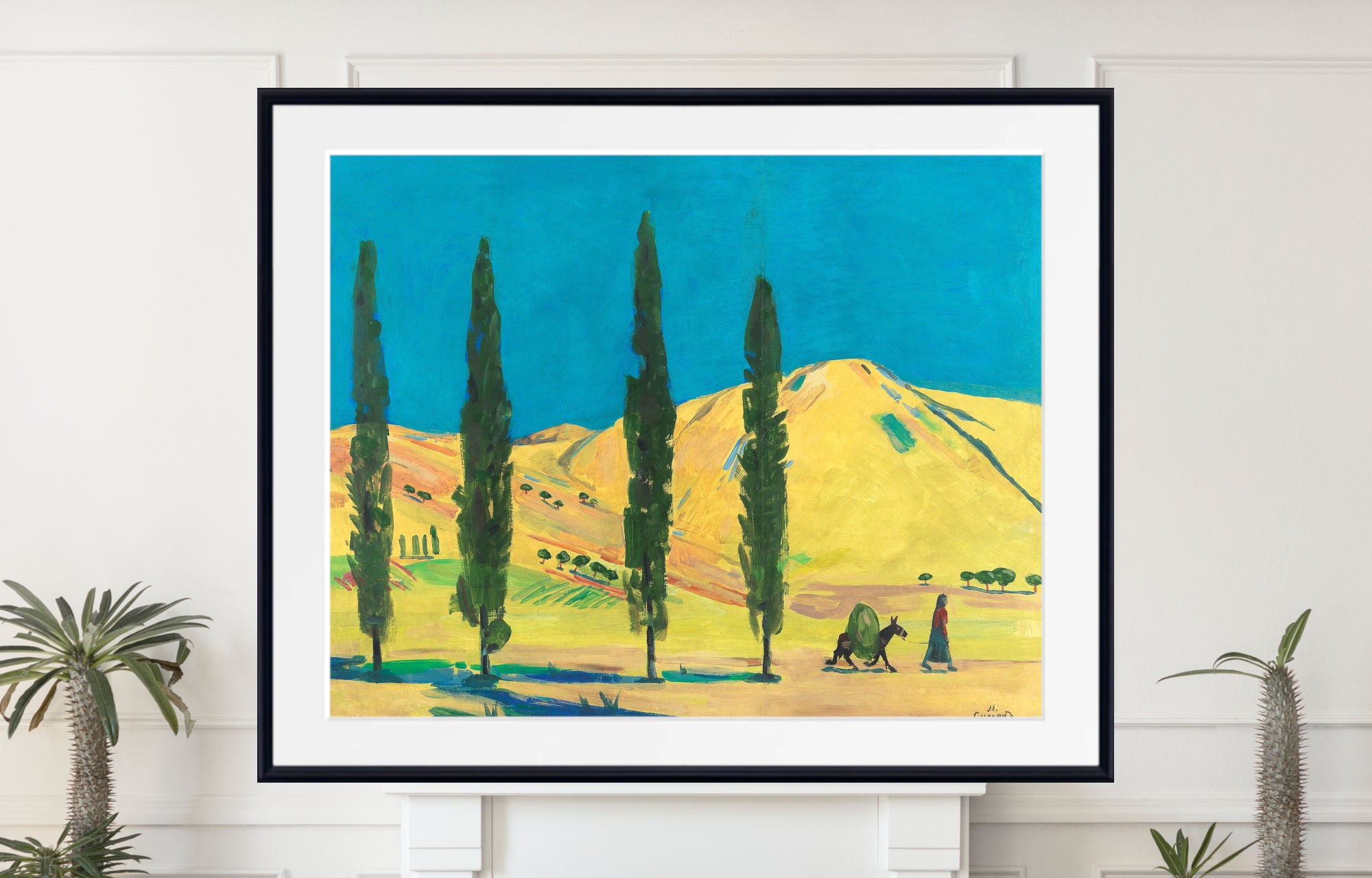 A framed art print depicting a vibrant landscape with tall cypress trees, rolling hills, and two camels under a bright blue sky, displayed on a white wall above a mantel with potted plants.