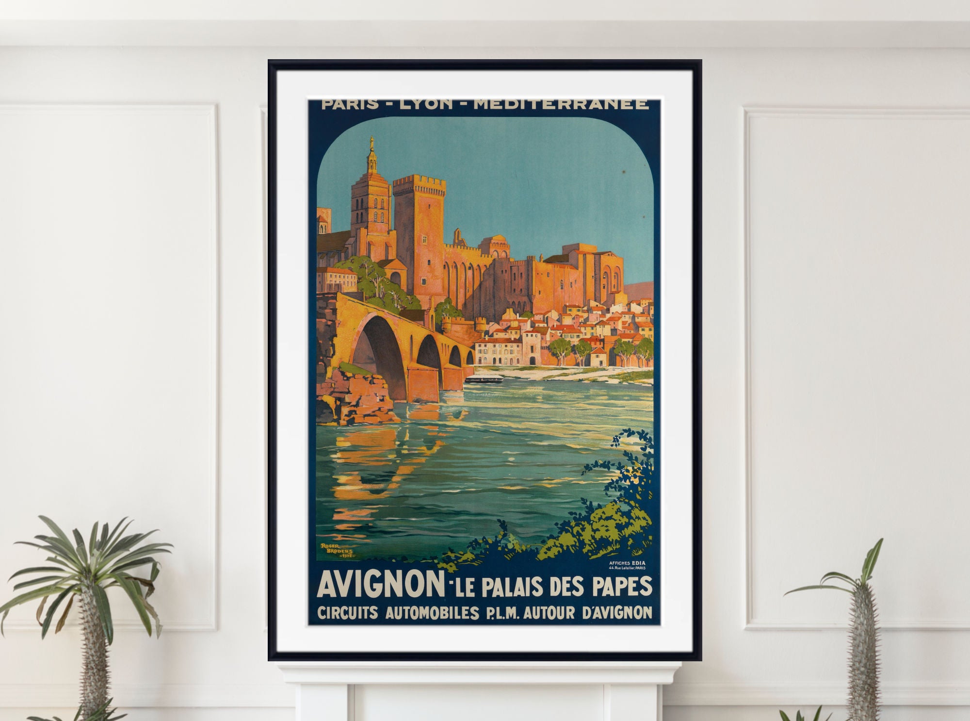 A framed vintage-style travel poster of Avignon, France, hanging on a white wall above a white mantel, with small potted plants at the sides. The poster depicts a cityscape with a bridge over water and the text 'AVIGNON - LE PALAIS DES PAPES'.