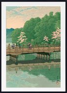 A framed Japanese-style art print depicting a scenic landscape with a wooden bridge over calm water, green trees, pink blossoms, and a pale sky. The print is shown with a white mat and a black frame.