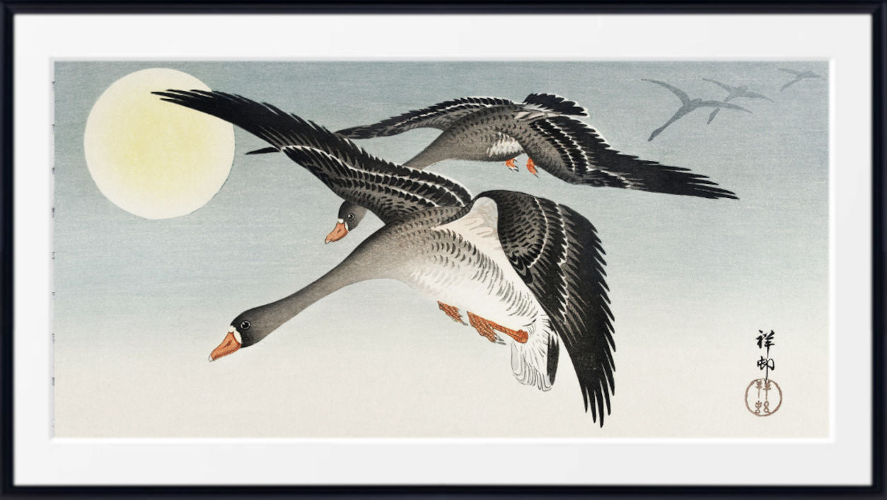 A framed Japanese-style art print depicting two flying geese with outstretched wings against a pale sky and large moon, shown in a black frame with white matting.
