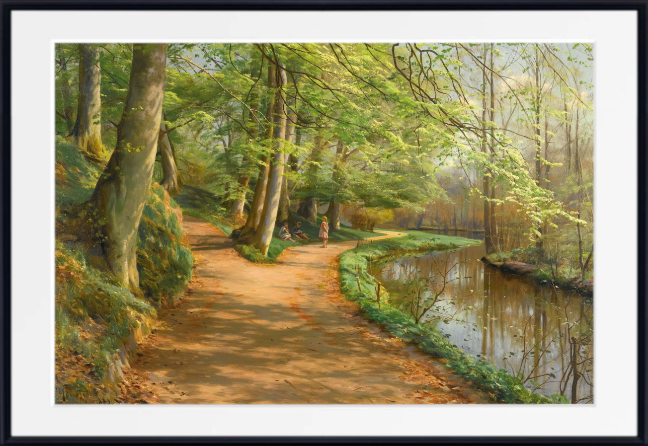 A framed landscape art print depicting a sunlit forest path running alongside a calm river, with tall trees, green foliage, and warm golden light. The artwork is presented with a wide white mat and a black frame.