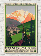 A vintage-style travel poster for Combloux, featuring a mountain landscape with snow-capped peaks, green hills, and a small town with a church spire, displayed against a white brick wall.