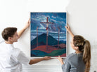 A man and a woman holding a framed art print depicting a dramatic landscape with a crucifix scene under a stormy blue sky.