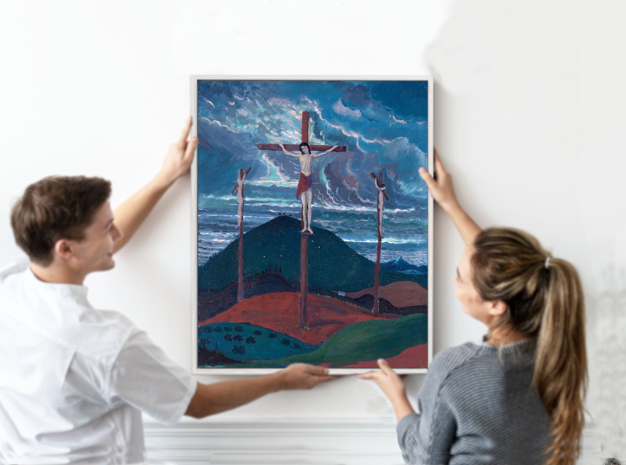 A man and a woman holding a framed art print depicting a dramatic landscape with a crucifix scene under a stormy blue sky.