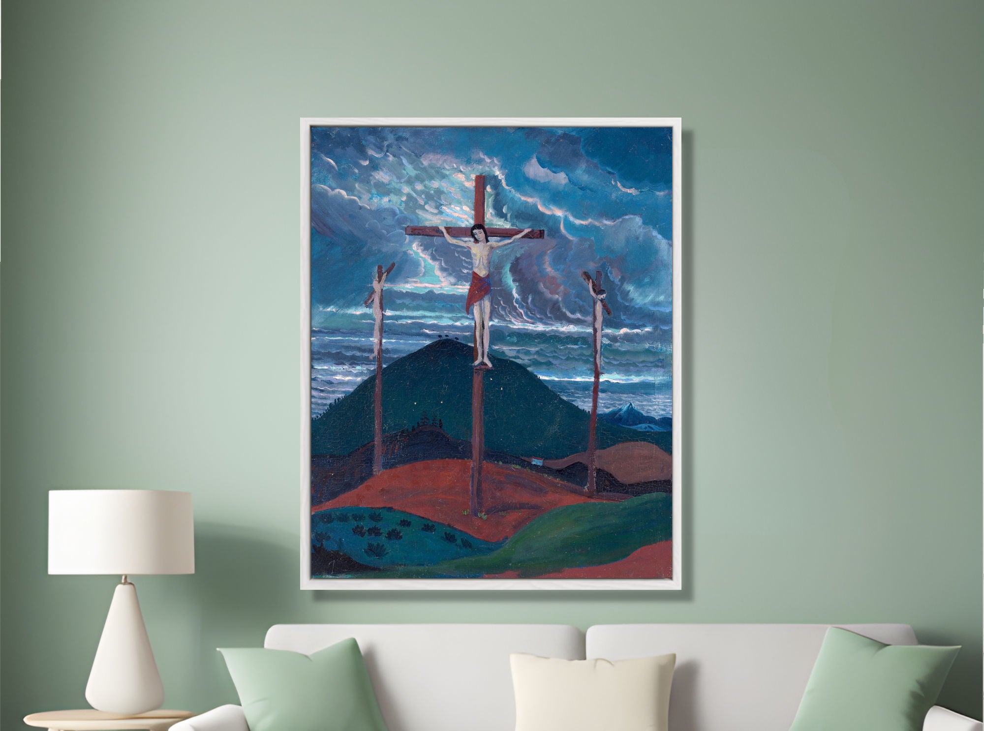 A framed art print depicting the crucifixion scene with three crosses against a stormy blue sky, displayed on a sage green wall above a white sofa with mint and cream pillows.