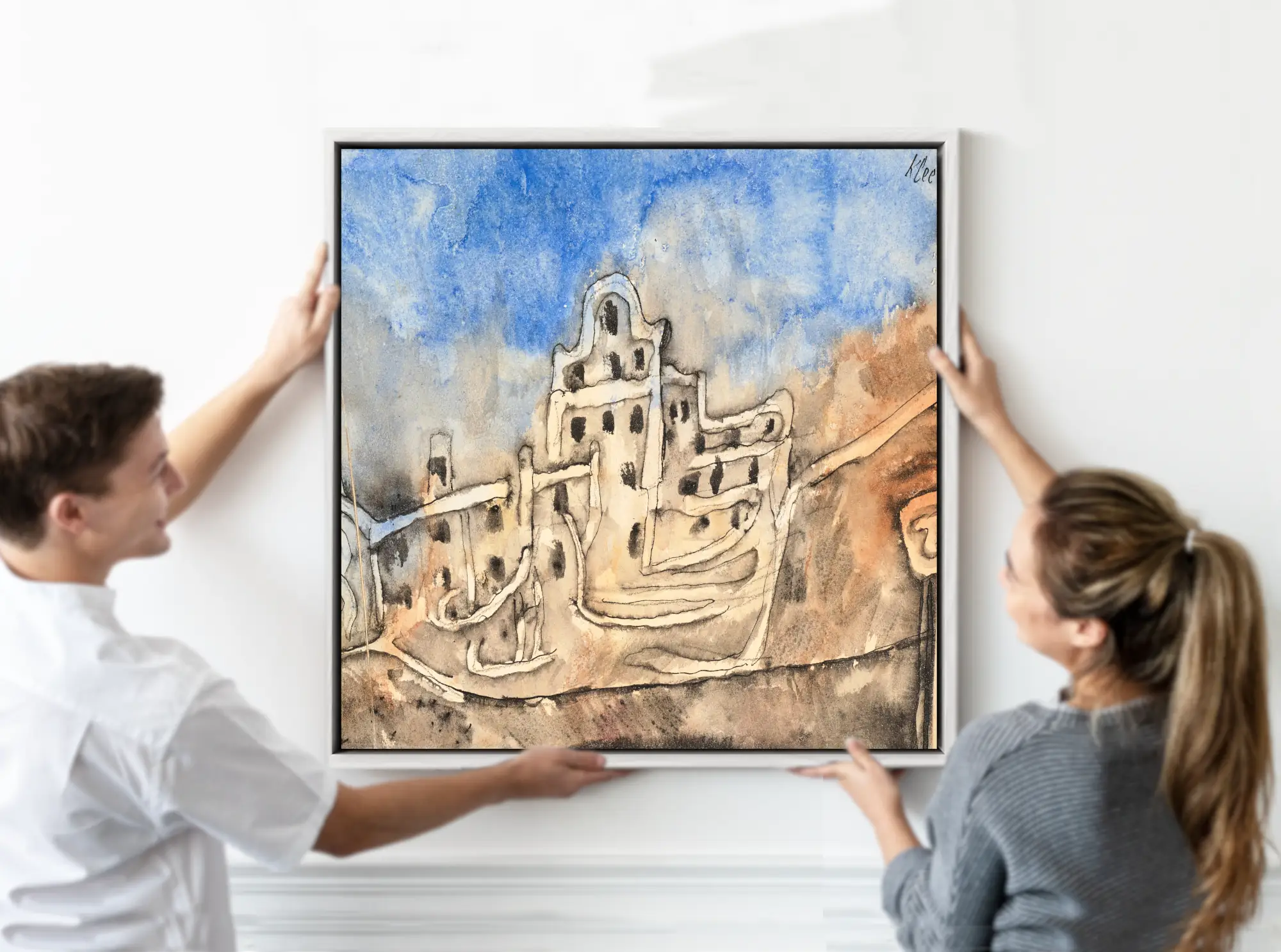 A framed watercolor painting of a hillside village with beige buildings under a blue sky, being held up by a man on the left and a woman on the right against a white wall. Paul Klee Fine Art Print, Der Palast (1912)