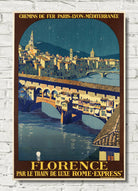 A vintage-style travel poster for Florence, Italy, featuring the Ponte Vecchio bridge over the Arno River at night, with the text 'CHEMINS DE FER PARIS-LYON-MÉDITERRANÉE' at the top and 'FLORENCE PAR LE TRAIN DE LUXE ROME-EXPRESS' at the bottom. The poster is displayed against a white brick wall background.