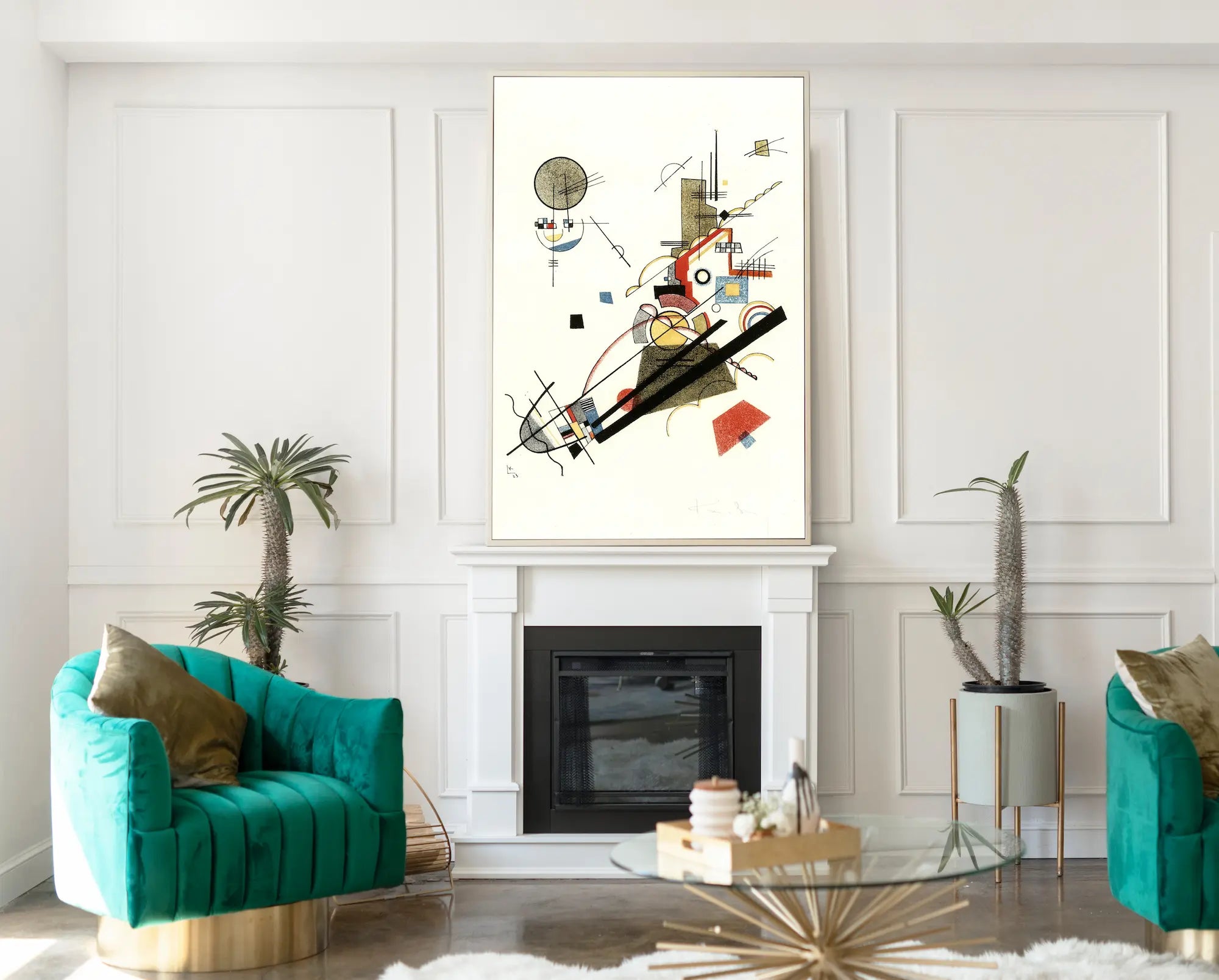 A framed abstract geometric wall art print hangs above a fireplace in a modern living room setting with teal velvet armchairs, potted plants, and a glass coffee table.