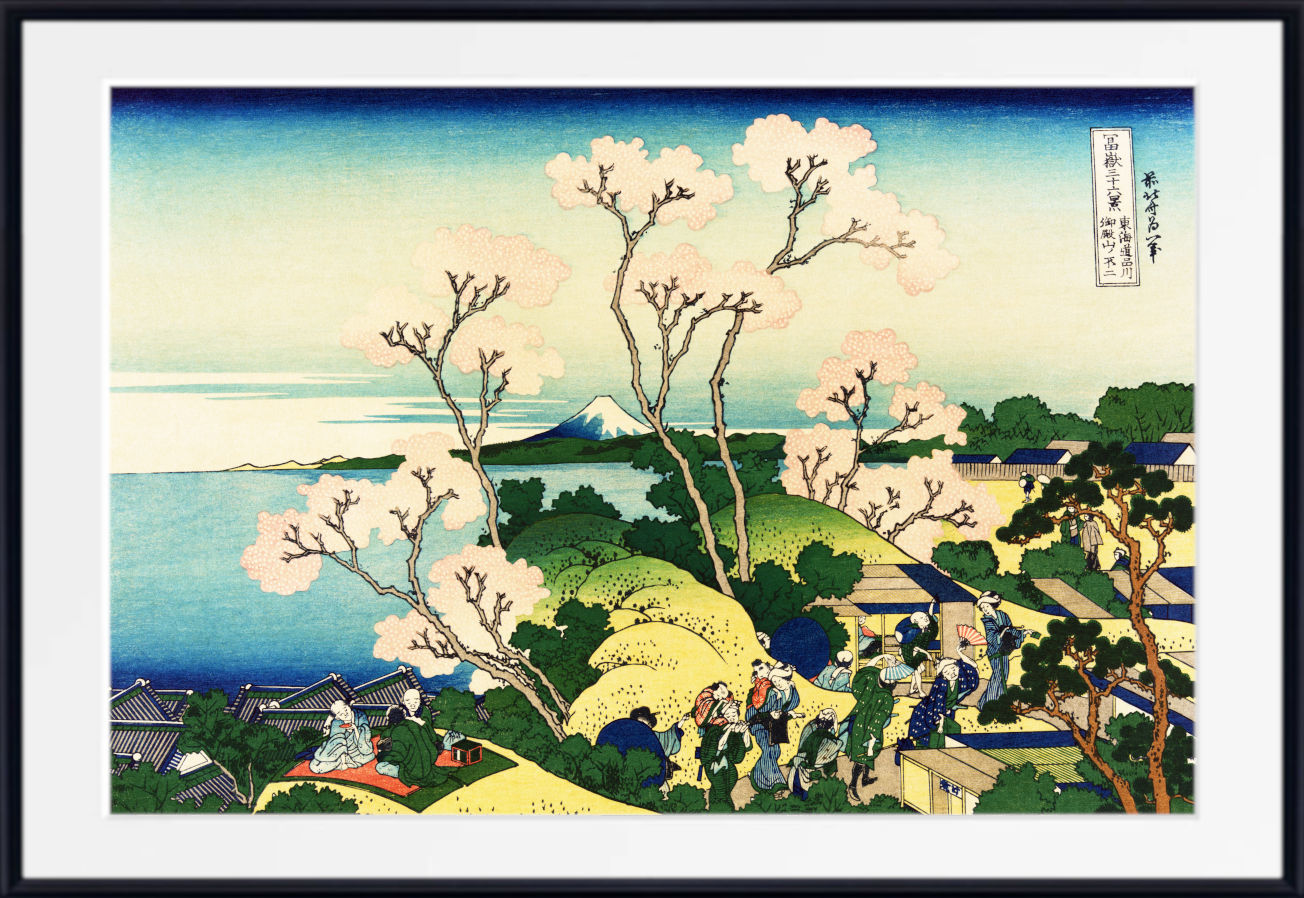 A framed Japanese ukiyo-e woodblock print depicting a coastal landscape with Mount Fuji, flowering trees, figures walking along a path, and buildings near the water. The print is shown with a white mat and a black frame against a light gray background.