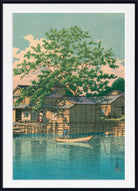 A framed Japanese-style art print depicting a serene lakeside village scene with wooden houses, a large green tree, and a small boat on blue water under a pale sky.