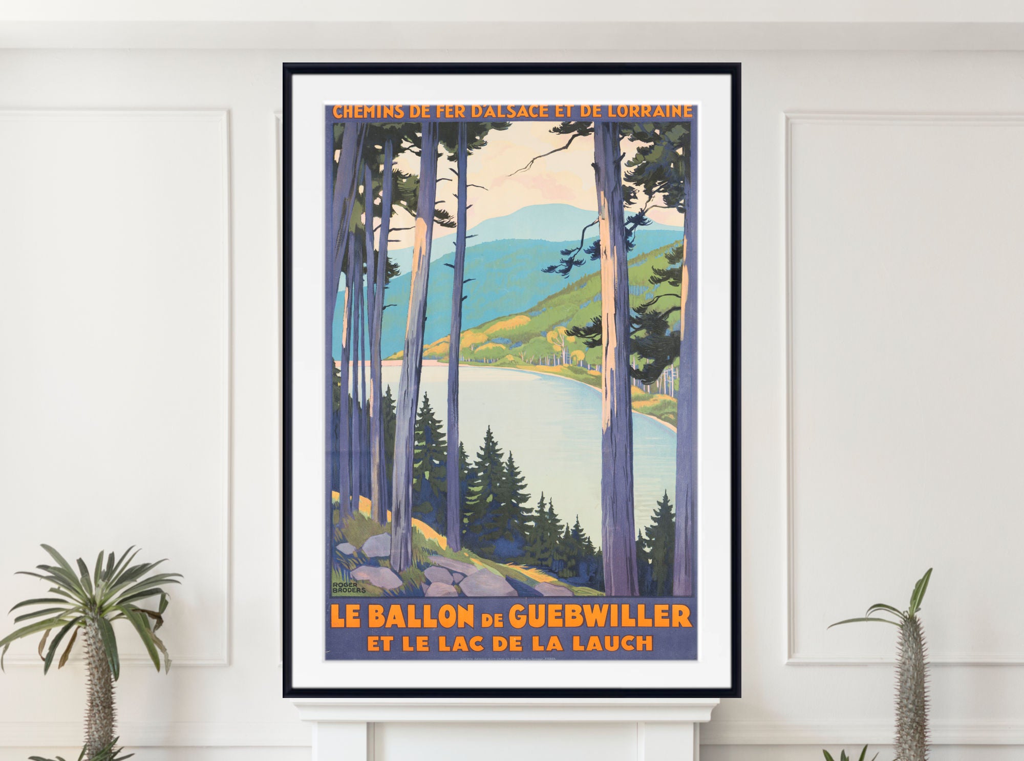 A framed vintage-style travel poster hangs on a white wall above a white mantel. The poster depicts a scenic mountain lake landscape with tall trees, blue mountains, green hills, and orange text reading 'Le Ballon de Guebwiller et le Lac de La Lauch.