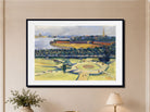 A framed landscape art print featuring a green park with a circular garden, water, and distant buildings, displayed on a beige wall above a small plant and lamp.