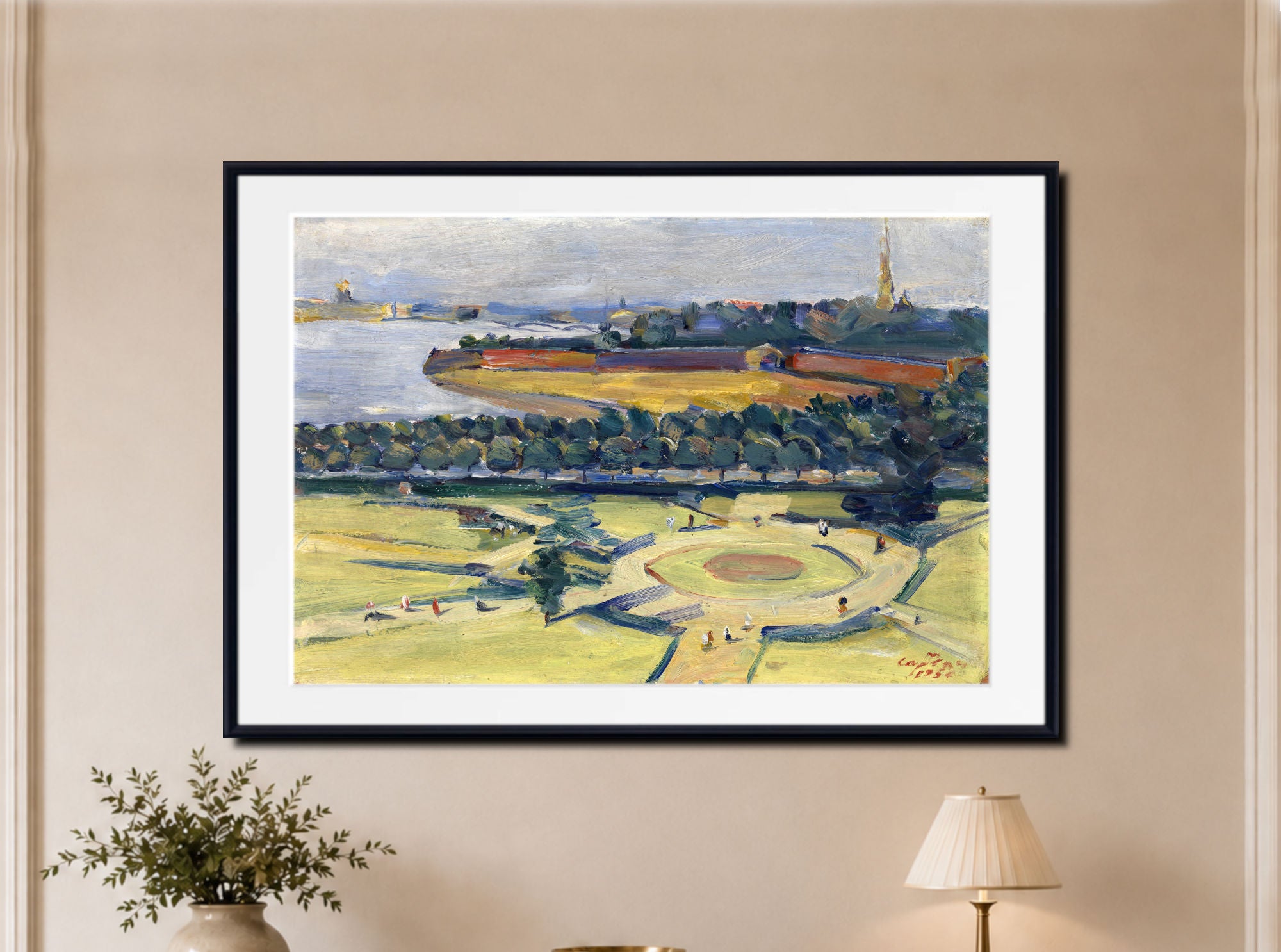 A framed landscape art print featuring a green park with a circular garden, water, and distant buildings, displayed on a beige wall above a small plant and lamp.