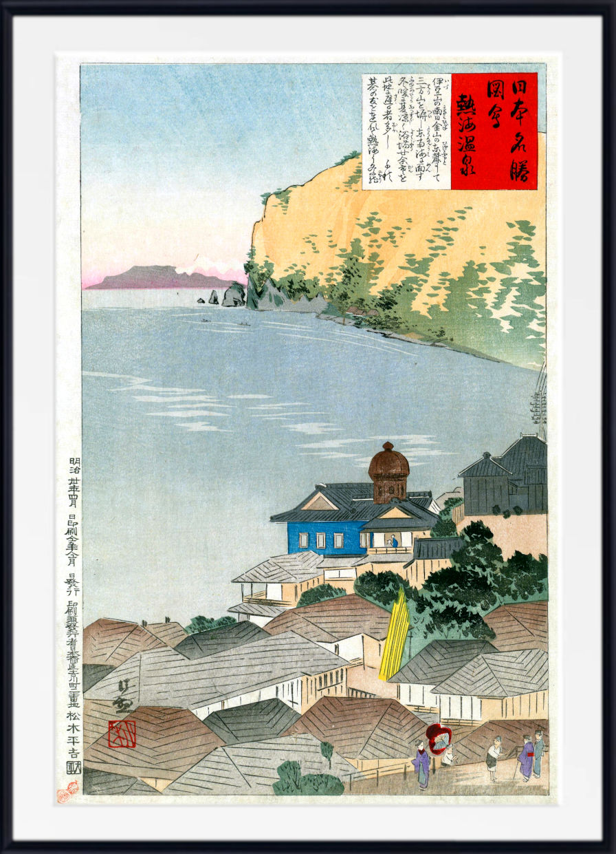 A framed Japanese ukiyo-e woodblock print depicting a coastal town with rooftops, a blue body of water, a pale yellow cliff, and a red title panel with Japanese text.