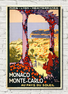 Vintage-style travel poster for Monaco and Monte-Carlo, featuring an illustrated woman in a red dress standing beneath a floral archway with a coastal cityscape in the background. The poster is displayed against a white brick wall.
