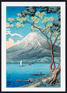 A framed Japanese-style art print depicting Mount Fuji over a blue lake, with a flowering tree in the foreground, small boats, and a white mat and black frame.