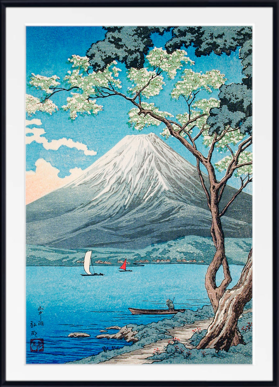 A framed Japanese-style art print depicting Mount Fuji over a blue lake, with a flowering tree in the foreground, small boats, and a white mat and black frame.